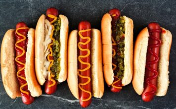 Dünya mutfağında geniş bir yer edinen hot dog, özellikle Amerika Birleşik Devletleri’nde adeta bir sembol haline gelmiştir. Kökeni 19. yüzyılın sonlarına kadar uzanan bu yiyecek, Almanya’dan Amerika’ya göç edenlerin beraberinde getirdiği sosis kültürü ile ortaya çıkmıştır. Uzun ekmek arasına yerleştirilen sosisin seneler içinde farklı soslarla buluşması, hot dog’u bugünkü popüler formuna kavuşturmuştur. Konulu bir haber görseli.
