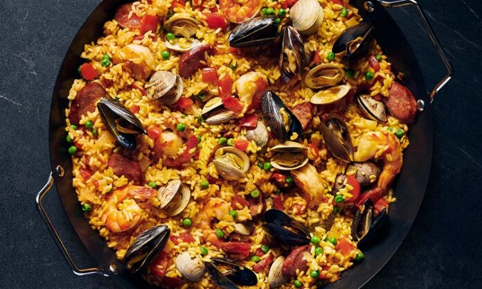 Paella, İspanya’nın en bilinen yemeklerinden biri olarak dünya mutfağında önemli bir yere sahiptir. Özellikle Valencia bölgesinde ortaya çıkan bu yemek, zamanla tüm ülkeye ve oradan da dünyaya yayılmıştır. Tarihi 15. yüzyıla kadar uzanan paella, köylülerin tarlalarda buldukları malzemeleri bir araya getirerek pişirdikleri pratik bir yemek olarak doğmuştur. Konulu bir haber görseli.