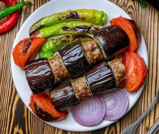 Türk mutfağının vazgeçilmez lezzetlerinden biri olan patlıcan kebabı, özellikle Gaziantep, Şanlıurfa ve Adana gibi şehirlerde sıkça hazırlanır. Patlıcanın közlenip etle buluşmasıyla ortaya çıkan bu yemek, hem hafif hem de doyurucu bir öğün sunar. Osmanlı mutfağından günümüze uzanan tarif, yöresel farklılıklarla her sofrada kendine özgü bir tat bırakır. Konulu bir haber görseli.