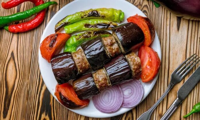 Türk mutfağının vazgeçilmez lezzetlerinden biri olan patlıcan kebabı, özellikle Gaziantep, Şanlıurfa ve Adana gibi şehirlerde sıkça hazırlanır. Patlıcanın közlenip etle buluşmasıyla ortaya çıkan bu yemek, hem hafif hem de doyurucu bir öğün sunar. Osmanlı mutfağından günümüze uzanan tarif, yöresel farklılıklarla her sofrada kendine özgü bir tat bırakır. Konulu bir haber görseli.